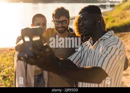 Eine vielfältige Gruppe von College-Freunden, die lustige Selfies mit einer Vintage-Sofortkamera machen und sich in der Nähe des Flusses bei Sonnenuntergang im Sommer entspannen. Hochwertige Fotos Stockfoto