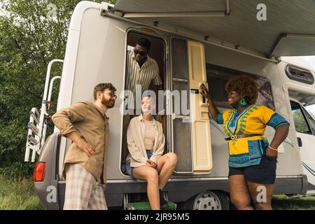 Campingurlaub im Sommer. Eine multikulturelle Gruppe von vier guten Freunden steht und redet, lacht und genießt es, Zeit zusammen in der Natur zu verbringen, während sie in ihrem komfortablen Wohnmobil leben. Hochwertige Fotos Stockfoto