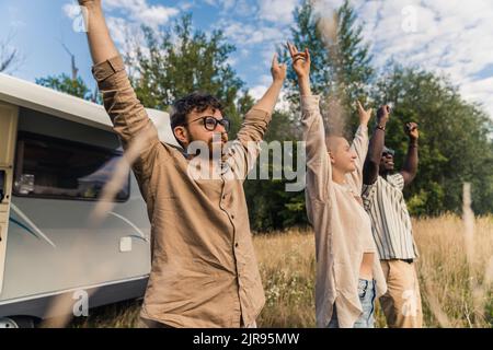 Eine Gruppe glücklicher, unbeschwerter bester Freunde, die in der Nähe ihres Wohnmobils stehen, die Hände heben und ein Friedenszeichen zeigen. Camping van Road Trip. Hochwertige Fotos Stockfoto