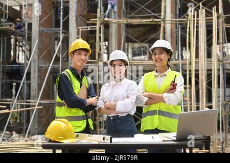 Bauingenieure und Spezialisten mit Harthüten und reflektierenden Jacken stehen mit gekreuzten Armen auf der Baustelle des Geschäftsgebäudes Stockfoto