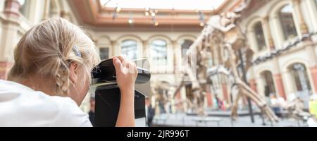 16.. Juni 2022- Berlin, Deutschland: Niedliches kleines entzückendes, neugieriges blondes Mädchen, das AR Fernglas Dinosaurier Skelett Ausstellung Naturgeschichte beobachtet Stockfoto