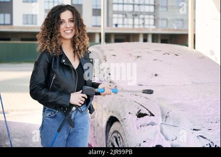 Fröhliche junge Frau in Lederjacke, die die Kamera anschaut und lächelt, während sie das Waschwerkzeug hält. Schöne stilvolle Frau mit Auto-Reinigungspistole stehen von Automobil mit Schaum bedeckt. Stockfoto
