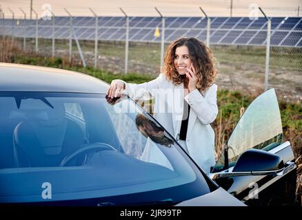 Mädchen, das am Telefon telefoniert, während sie in der Nähe eines offenen Autos steht und nach links schaut. Auf einem Backgdrop-Solarkraftwerk, das nachhaltigen Strom und erneuerbare Energie erzeugt. Wirtschaftliche und ökologische Energie indystry. Stockfoto
