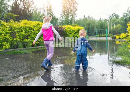 Rückansicht von zwei niedlichen blonden kaukasischen wenig Kinder Bruder und Schwester genießen Spaß haben spielen springen in Schmutzige Pfütze in Blau Stockfoto