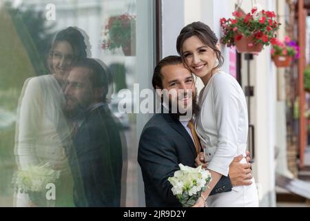 Schönes Hochzeitspaar, das während des Hochzeitsspaziergangs neben einem großen Glasfenster sitzt. Das Brautpaar lächelt und schaut auf die Kamera Stockfoto