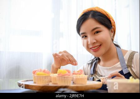 Schöne junge asiatische weibliche Bäcker dekorieren ihre schöne hausgemachte Cupcake mit winzigen weißen Schokoladenkugel. Stockfoto