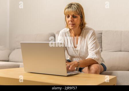 Reife Frau, die zu Hause auf dem Sofa sitzt und auf ihrem Laptop auf einem kleinen Holztisch im Wohnzimmer tippt. Stockfoto