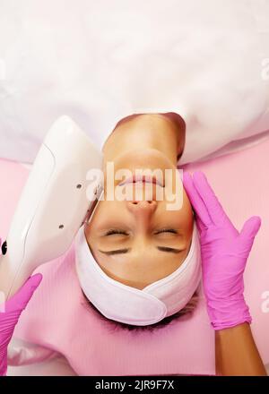 Draufsicht auf junge Frau, die mit geschlossenen Augen liegt und mit einem Laser-Gerät für Schönheitsgeräte eine Verjüngungsbehandlung erhält. Stockfoto