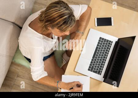 Frau, die von zu Hause aus mit ihrem Laptop und Mobiltelefon neben ihr in ihr Tagebuch schreibt. Aufnahme über Kopf Stockfoto