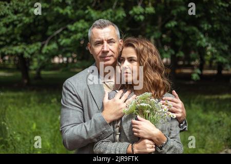 Fröhliche Familie, die im Sommer im Park steht. Selbstbewusster älterer Mann umarmt junge Frau mit einem Bouquet von Kamillen. Stockfoto