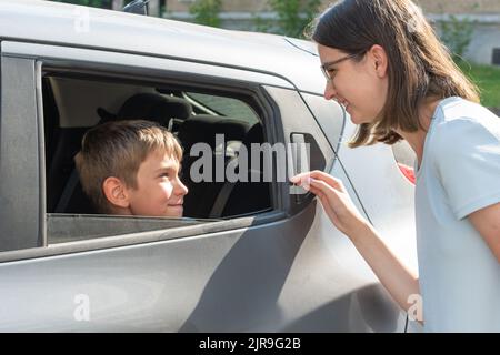 Mama spricht mit ihrem Sohn durch das Autofenster und wünscht ihm viel Erfolg in der Schule. Glückliches Kind, das im Auto sitzt und mit Mutter spricht. Das Konzept der Rückkehr zu Stockfoto