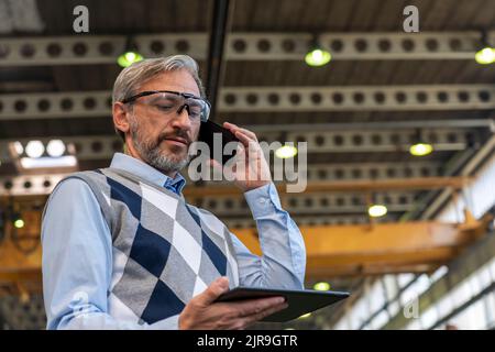 Taille nach oben Porträt eines selbstbewussten, eleganten, grauhaarigen CEO oder Geschäftsmanns. Mann Steht Und Benutzt Smartphone Und Digitales Tablet. Stockfoto