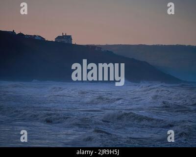 Wütende Sturmwellen stürzen in die Bucht zwischen Whitby und Sandsend an der Nordseeküste, während die Wintersonne hinter der Landzunge und den Klippen untergeht. Stockfoto