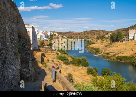 Der Fluss Guadiana fließt durch das historische Burgdorf Mertola mit der Brücke Ponte de Mertola im Hintergrund. Stockfoto