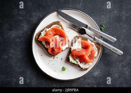 Roggenbrot mit Frischkäse und geräuchertem Lachs Toast auf Teller, schwarzer Stein Hintergrund. Leckerer Toast mit rotem Fisch oder offenes Sandwich Stockfoto