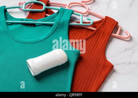 Solides Deodorant auf blaugrünem und rotem Tanktop auf Marmorboden. Antitranspirant-Stick auf einem Leinen-Jersey Tank-Shirts auf Kleiderbügel. Körperpflege, Hautpflege. Stockfoto