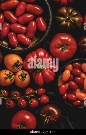 Frische, rohe und bunte Tomaten in verschiedenen Größen und Arten auf dunklem Hintergrund Stockfoto