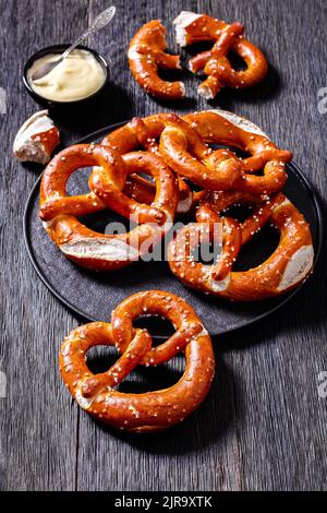 Weiche Brezeln in Form von Knoten gebacken und mit Salz auf schwarzem Teller auf dunklem Holztisch mit Käsesauce bestreut, vertikale Ansicht von oben Stockfoto