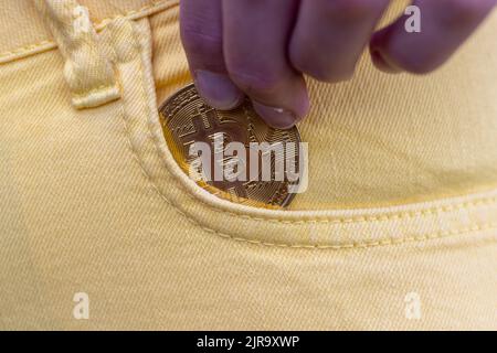 Nahaufnahme einer Frau, die Bitcoin in die gelbe Jeanstasche steckte, goldene Kryptowährung-Münze im Taschenhintergrund, Konzept der digitalen Währung virtuelles Geld. Hochwertige Fotos Stockfoto