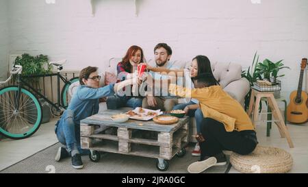Glückliche Freunde haben Party reden und Pizza essen. Sie klirken Flaschen, die zuhause auf dem Sofa sitzen Stockfoto