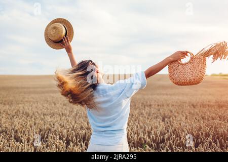 Rückansicht der jungen glücklichen Frau springen genießen Blick im Sommerfeld hält Stroh Handtasche mit Weizen Bundle. Stockfoto