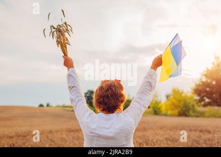 Unabhängigkeitstag der Ukraine. Glückliche Frau hob die Arme mit blauen und gelben Fahnen auf dem Feld mit einem Weizenbündel Stockfoto