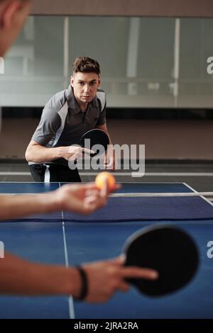 Ernsthafter junger Mann, der mit einem Freund im Fitnessstudio Tischtennis spielt, wenn er für den Wettkampf trainiert Stockfoto