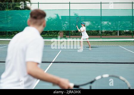 Lächelnde junge Frau, die beim Tennisspielen mit einem Freund Ball serviert Stockfoto