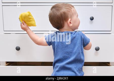 Kleinkind Baby öffnet eine Kommode. Der Junge greift in eine offene Schublade eines weißen Schranks. Kind im Alter von einem Jahr Stockfoto