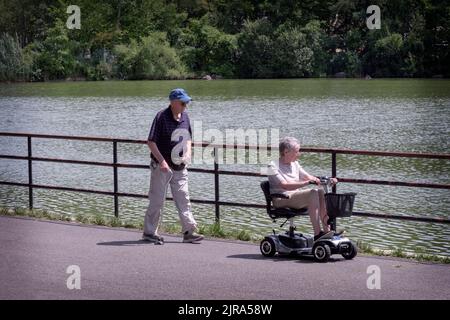 Ein Paar mit gesundheitlichen Problemen, vermutlich verheiratet, teilt sich einen Spaziergang/eine Fahrt um den See im Kissena Park, Flushing, New York City. Stockfoto