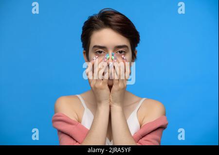 Skinny niedlichen Teenager-Mädchen versteckt Untergesicht in ihren Händen, blauer Hintergrund Stockfoto