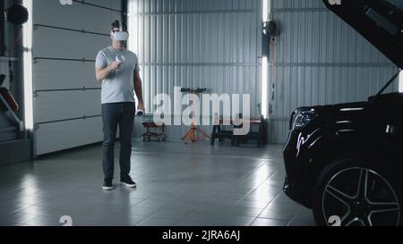 In einem Autodienst ein Mann Mechaniker in Virtual-Reality-Brille mit Steuergeräten bewegen seine Hände während der Diagnose und Inspektion eines Autos mit geöffneter Haube Stockfoto
