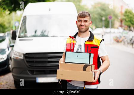 Die Lieferungen sind termingerecht. Porträt eines lächelnden Lieferers, der vor seinem Lieferwagen steht und ein Paket hält. Stockfoto