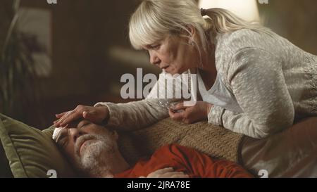 Kippen Sie die Ansicht nach unten, dass eine ältere Frau auf dem Sofa zu Hause eine Tablette gab und die Stirn eines kranken älteren Mannes berührte, der an Fieberschmerzen litt Stockfoto