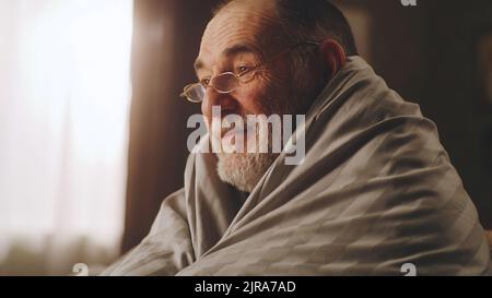 Älterer Mann, der während der Gaskrise mit einer warmen Decke bedeckt war und einen Film im Fernsehen sah, während er auf der Couch im Wohnzimmer saß Stockfoto