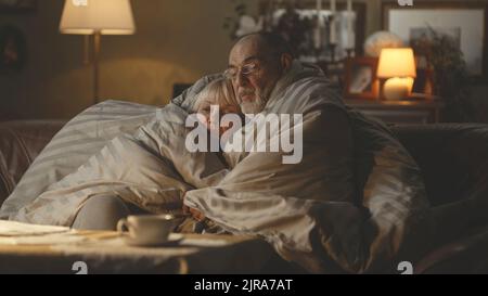 Verärgert Senior Mann und Frau umarmen einander und sprechen unter warmen Bettdecke, während auf dem Sofa sitzen in kalten Wohnung während der Gas-Krise Stockfoto