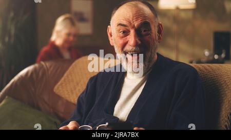 Überraschte ältere Männer, die sich die Brille ausnahmen und staunend auf die Kamera schauten, nachdem sie gute Nachrichten auf dem Handy gelesen hatten, während sie auf dem Sofa im Wohnzimmer zu Hause saßen Stockfoto