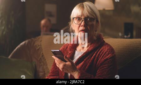 Verängstigte Frau in einer Brille, die mit geöffnetem Mund auf die Kamera schaute, während sie auf der Couch saß, nachdem sie am Abend zu Hause schockierende Nachrichten auf dem Handy gelesen hatte Stockfoto
