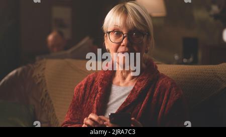 Eine verwunschte ältere Frau, die sich die Brille auszieht und lächelnd auf die Kamera schaut, nachdem sie auf dem Smartphone angenehme Nachrichten gelesen hat, während sie im dunklen Wohnzimmer zu Hause auf der Couch sitzt Stockfoto