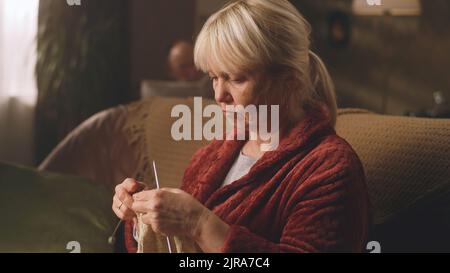 Eine alte Großmutter in einem Bademantel, die einen warmen Pullover mit Stricknadeln kreiert, der auf dem Sofa in einem gemütlichen Wohnzimmer sitzt Stockfoto