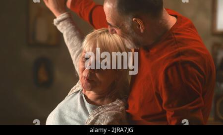 Neigen Sie die Ansicht eines älteren Mannes, der sich küsst und eine gealterte Frau umarmt, während er im Wohnzimmer zu Hause zusammen tanzt Stockfoto