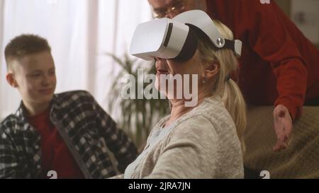 Fröhliche Großmutter in Virtual-Reality-Brille auf Sofa lachend und gestikulend in der Nähe glücklicher Enkel und Großvater im Wohnzimmer Stockfoto
