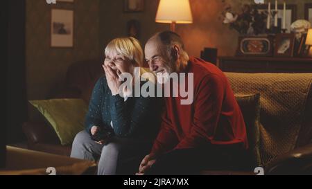 Glücklicher älterer Mann und Frau, die über einen lustigen Witz lachen, während sie zu Hause auf der Couch sitzen und abends Comedy im Fernsehen schauen Stockfoto