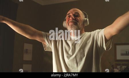 Niedriger Winkel von glücklichen älteren Männern in weißem T-Shirt und kabellosen Kopfhörern, Musik hören und Arme schwingen während des Fitnesstrainings am Morgen zu Hause Stockfoto