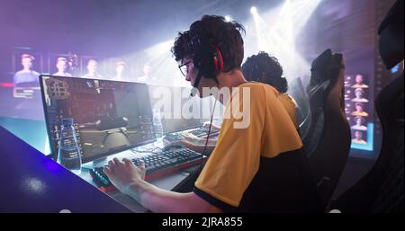 Esportsman Gamer in Brille und Kopfhörer spielen Shooter auf Computer in der Nähe des Teams, während Park in der Profi-Gaming-Meisterschaft Kampf Stockfoto