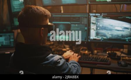 Junger Mann in VR-Brille, der Computer benutzt, um den Start einer Atombombe zu hacken, um Krieg zu beginnen, während er im dunklen Raum sitzt Stockfoto