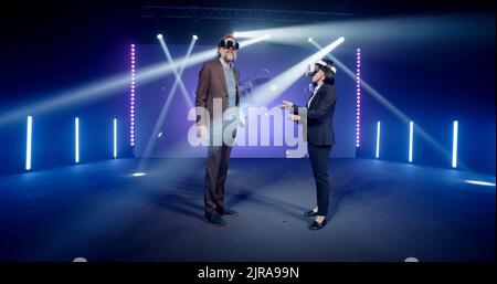 Zwei Moderatoren in Anzügen auf der Bühne, die neue Augmented-Reality-Brillen bei einer Metauniversum-Konferenz in einem Studio mit Scheinwerfern vor dem LED-Bildschirm mit 3D Objekten ausprobieren Stockfoto