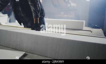 Nicht erkennbarer Mann in Freizeitkleidung und Handschuhen, der Plastikblock mit Klebeband misst, bevor er während der Arbeit in der professionellen Werkstatt Polymer auf die Werkbank setzt Stockfoto