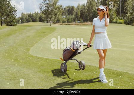 Frau Golfspielerin, die mit dem Golfwagen auf dem Fairway läuft. In weißer sportiver Uniform. Kurzer Rock, Polo und Mütze Stockfoto