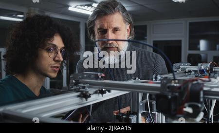 Männlicher Designer mit lockigen Haaren, der während der Arbeit im zeitgenössischen Studio die Arbeit des Druckers 3D einem reifen bärtigen Kollegen vorführt und erklärt Stockfoto